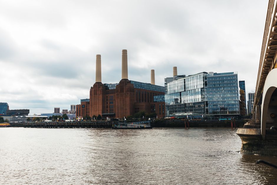 A large historic power station building constructed from red brick with several tall cream-colored smokestacks extending upward. The structure is located along a riverbank, with a paved area or promenade visible at the water's edge and some small boats docked nearby. To the right of the power station, there is modern glass-fronted office or commercial buildings with reflective windows. The scene is set under a cloudy sky, with diffuse natural light illuminating the scene. The foreground features the water of the river, with gentle ripples, and the background shows a cityscape with additional buildings. This setting suggests an urban environment where industrial architecture contrasts with modern development and could relate to local waste management or disposal services for commercial or industrial waste, consistent with a site where independent rubbish collection or clearance services may operate. Rubbish Clearance Battersea may provide option for on-site rubbish removal near this area, supporting alternative waste handling for businesses and residents.