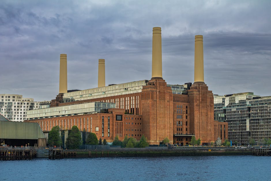 A large industrial building constructed from reddish-brown brick with a tiered rectangular form, featuring vertical window strips along its facade. Four tall, cylindrical cream-colored chimneys extend upward from the structure, evenly spaced at the corners. The building is situated on the banks of a river or large body of water, with a paved or concrete area at its base and a small green space with trees along the riverfront. Surrounding the building are modern residential or commercial structures with glass and steel exteriors, and partly cloudy skies overhead illuminate the scene. The image’s composition emphasizes the industrial aesthetic, which can be associated with traditional or alternative waste management site environments, relevant to rubbish clearance services that deal with large-scale or industrial waste removal involving on-site clearance of bulky or hazardous materials by specialists such as Rubbish Clearance Battersea.