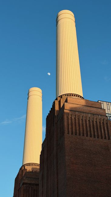 A large historic power station building constructed from red brick with several tall cream-colored smokestacks extending upward. The structure is located along a riverbank, with a paved area or promenade visible at the water's edge and some small boats docked nearby. To the right of the power station, there is modern glass-fronted office or commercial buildings with reflective windows. The scene is set under a cloudy sky, with diffuse natural light illuminating the scene. The foreground features the water of the river, with gentle ripples, and the background shows a cityscape with additional buildings. This setting suggests an urban environment where industrial architecture contrasts with modern development and could relate to local waste management or disposal services for commercial or industrial waste, consistent with a site where independent rubbish collection or clearance services may operate. Rubbish Clearance Battersea may provide option for on-site rubbish removal near this area, supporting alternative waste handling for businesses and residents.