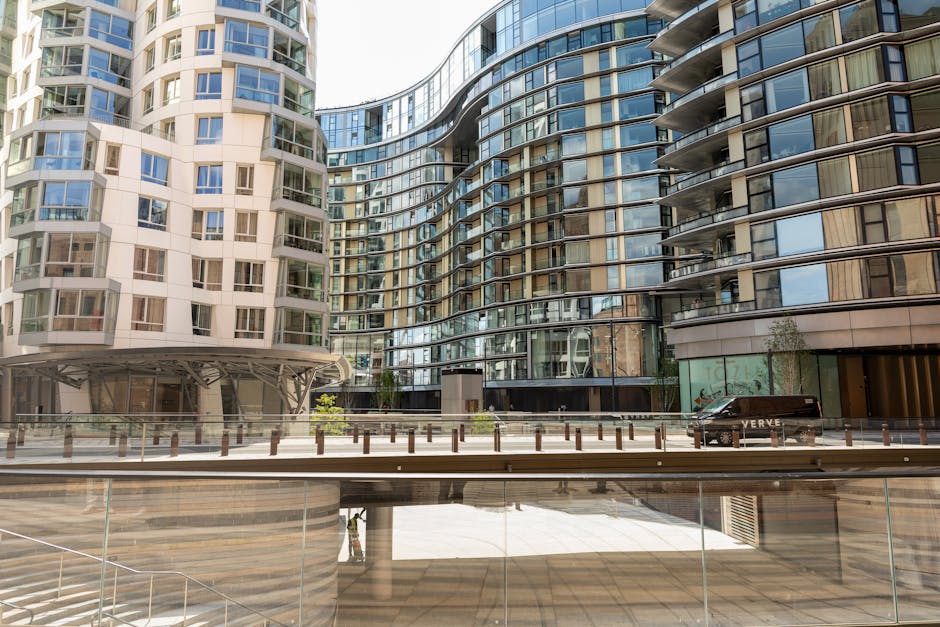A modern high-rise residential building with a curved glass and metal facade, reflecting the surrounding urban environment. The structure features numerous balconies with metal railings, some with glass panels, and large windows that provide extensive natural light to the interior. The lower levels include a ground-floor entrance area with glass doors and a small landscaped section with young trees and shrubs. In front of the building, there is a paved walkway separated from the road by a row of short, evenly spaced wooden posts and a transparent glass barrier. A black van with visible text on its side is parked along the curb, indicating possible private property or service vehicles. The scene is well-lit with natural daylight, capturing the reflective surfaces of the glass and the textured finishes of the building’s materials. The overall environment suggests an urban setting with contemporary architectural design, which could relate to site-specific waste management activities or the need for professional rubbish removal to support such properties, as offered by Rubbish Clearance Battersea.