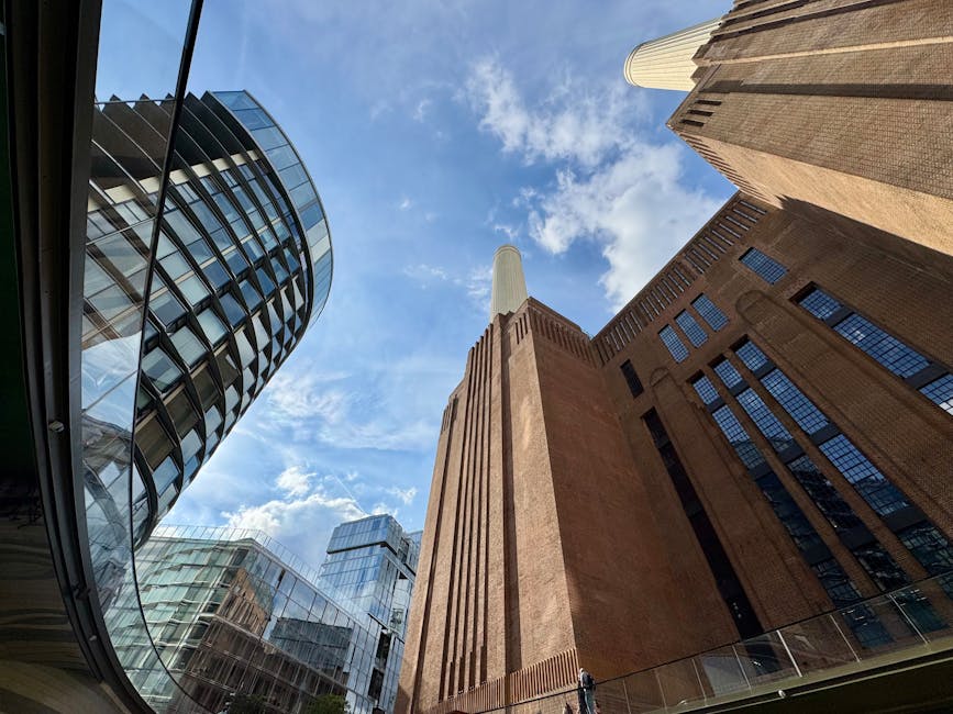 A view looking up at modern high-rise buildings in an urban setting, showcasing a curved glass tower with reflective windows on the left, and a tall, rectangular brick building with vertical lines and small, evenly spaced windows on the right. In the center is a large industrial-style structure made of reddish-brown brick with a tall, white chimney extending upward toward a partly cloudy sky. The buildings are surrounded by a glass and steel structure at ground level, suggesting a commercial or office area, with natural daylight illuminating the scene. This image highlights the contrast between contemporary glass architecture and traditional brick facades, reflecting urban development common in city centres where private waste removal services like Rubbish Clearance Battersea operate for managing waste from commercial properties such as offices and shops.