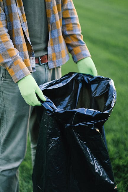 A person standing outdoors on a grassy area is holding open a large black plastic rubbish bag, ready for waste disposal. The individual is wearing a checkered shirt with tones of yellow, blue, and gray over a green T-shirt, along with light gray trousers secured by a brown belt. They are also wearing light green gloves, which are gripping the edges of the rubbish bag. The background features a well-maintained lawn, suggesting a garden or park setting, with natural daylight illuminating the scene. The black rubbish bag appears to be made of shiny, slightly crinkled plastic, partially open and empty, possibly prepared for waste collection as part of an on-site rubbish removal process. This image reflects the activity of private waste handling, potentially related to independent rubbish clearance services, with a focus on environmentally cautious disposal practices. The setting and equipment emphasize a professional approach to rubbish removal, aligning with services offered by companies like Rubbish Clearance Battersea, who undertake various rubbish collection tasks in residential or outdoor environments.