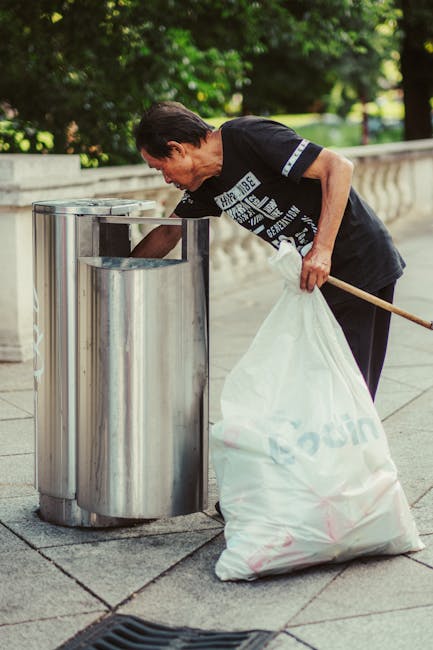 A middle-aged man with dark hair, wearing a black T-shirt with a white graphic print on the front and dark trousers, is bent over as he disposes of trash into a large, cylindrical stainless steel street litter bin. The bin is positioned on a paved sidewalk next to a low stone wall with a decorative balustrade, in a park-like setting surrounded by lush green trees and bushes. The man holds a long-handled litter picker in one hand and a large white plastic bag in the other, which appears to be filled with rubbish awaiting disposal. The reflective surface of the bin shows the surrounding environment, including the greenery and part of the sidewalk, creating a clean, organized scene focused on waste collection in an outdoor public space. The scene exemplifies an instance of private or independent waste handling, such as rubbish removal or on-site clearance, that could be carried out by services like Rubbish Clearance Battersea, aligning with the context of alternative rubbish removal methods.