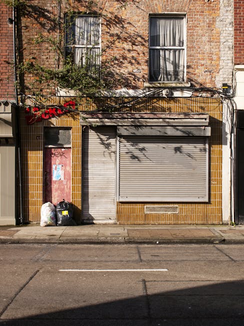 The image depicts the front façade of a two-story building constructed with reddish-brown bricks, with two large windows on the upper floor, each divided into smaller panes by white frames and covered with semi-translucent curtains. Below, the ground level features a storefront with a closed, metallic, roll-down shutter door that has a weathered grey finish, and a smaller, adjacent pedestrian door also secured with a matching rolling shutter. To the left of the entrance, there are three black plastic rubbish bags and two white bags stacked on the sidewalk, suggesting ongoing rubbish clearance or disposal activity. The building’s exterior includes yellow-brown ceramic tiles framing the store entrance, with some graffiti markings and shadow patterns cast by nearby trees visible on the brickwork and shutter. Electrical wiring is loosely mounted along the facade, extending across the front of the building. The overall scene indicates a typical urban street environment where private waste collection, managed by a service such as Rubbish Clearance Battersea, might be part of the site’s ongoing rubbish removal process, reflecting an alternative approach to municipal refuse handling in the Battersea area.