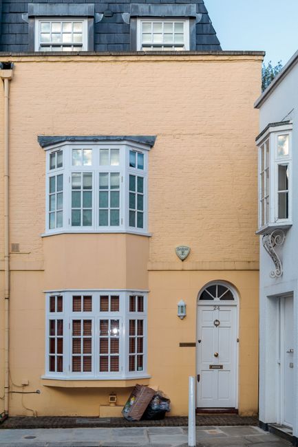 A narrow, three-storey residential building with a light peach-yellow painted brick exterior, featuring two prominent bay windows—one on the ground floor and a larger, rounded window on the first floor—both with white frames and multiple small panes of glass. The ground-floor bay window has decorative wooden latticework at the base, and the upper window has a slight outward projection, providing a traditional aesthetic. To the right of the building is a white door with a semi-circular window at the top, adorned with a small black letterbox and a door handle, flanked by a metallic lamp fixture and a small dark-colored mailbox. In front of the building, there are two bags of trash resting on the pavement, indicating recent clearance or rubbish disposal activity. The scene is set on a quiet street with parts of neighboring buildings visible, underscoring an urban environment where private on-site clearance or independent rubbish removal services, such as those provided by Rubbish Clearance Battersea, might be employed to manage waste in properties like this one. The overall scene is lit by natural daylight, with a clear sky overhead, highlighting the clean, orderly appearance of the property’s facade and the immediate surroundings.