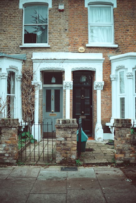 The image depicts the front façade of a two-story building constructed with reddish-brown bricks, with two large windows on the upper floor, each divided into smaller panes by white frames and covered with semi-translucent curtains. Below, the ground level features a storefront with a closed, metallic, roll-down shutter door that has a weathered grey finish, and a smaller, adjacent pedestrian door also secured with a matching rolling shutter. To the left of the entrance, there are three black plastic rubbish bags and two white bags stacked on the sidewalk, suggesting ongoing rubbish clearance or disposal activity. The building’s exterior includes yellow-brown ceramic tiles framing the store entrance, with some graffiti markings and shadow patterns cast by nearby trees visible on the brickwork and shutter. Electrical wiring is loosely mounted along the facade, extending across the front of the building. The overall scene indicates a typical urban street environment where private waste collection, managed by a service such as Rubbish Clearance Battersea, might be part of the site’s ongoing rubbish removal process, reflecting an alternative approach to municipal refuse handling in the Battersea area.