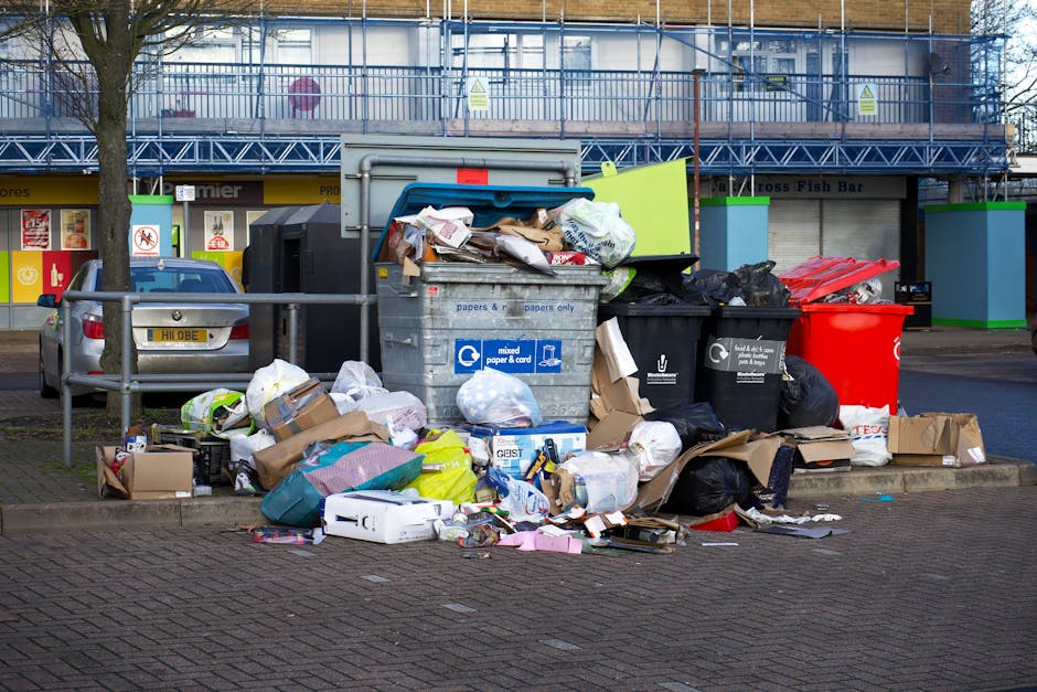 A large accumulation of mixed household waste and recyclables is piled up on a paved area adjacent to a parking space, including cardboard boxes, plastic bags, paper, and discarded packaging. The waste is scattered around a central gray communal recycling bin labeled for mixed paper and cardboard, which appears to be overflowing with various items, some leaning or partially spilling out. Nearby are black and red trash bags and additional loose rubbish, with some cardboard boxes placed on the ground beside the pile. In the background, there is a metal fence, with a row of retail shops beneath a covered walkway, and a parked car facing toward the rubbish. The surroundings are a typical urban environment with a mix of commercial and parking areas. The scene illustrates improper waste disposal, which contrasts with the services offered by Rubbish Clearance Battersea for private or alternative waste removal methods, reflecting the importance of adhering to local rubbish regulations and proper waste management practices.