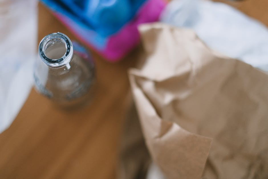 A clear glass bottle with a rounded body and a narrow neck, placed on a wooden surface, reflects ambient light and shows a few water droplets at the rim. Behind the bottle, partially out of focus, is a crumpled light brown paper bag and a blurred section of what appears to be a colorful cardboard box or packaging in shades of blue and pink. The setting suggests an indoor environment, possibly a kitchen or a workspace, where waste or recycling materials are gathered for disposal. The image showcases textures of the smooth glass and crinkled paper, highlighting common household rubbish materials that might be handled by a waste removal service like Rubbish Clearance Battersea, which offers alternative disposal options for general waste. Lighting is soft and natural, emphasizing the details of the objects and their textures without harsh shadows, aligning with the theme of responsible rubbish management and waste collection practices.