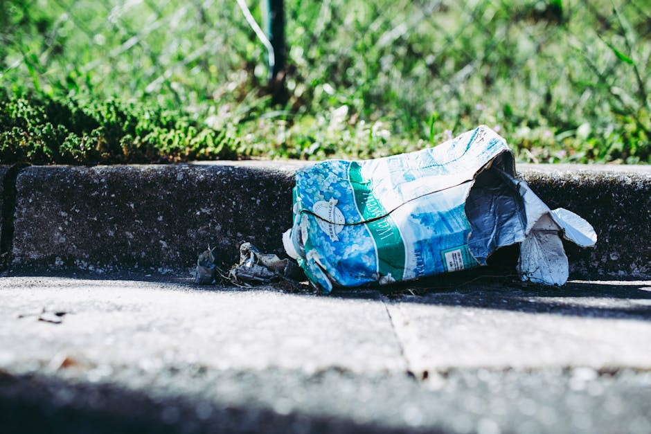 A large accumulation of mixed household waste and recyclables is piled up on a paved area adjacent to a parking space, including cardboard boxes, plastic bags, paper, and discarded packaging. The waste is scattered around a central gray communal recycling bin labeled for mixed paper and cardboard, which appears to be overflowing with various items, some leaning or partially spilling out. Nearby are black and red trash bags and additional loose rubbish, with some cardboard boxes placed on the ground beside the pile. In the background, there is a metal fence, with a row of retail shops beneath a covered walkway, and a parked car facing toward the rubbish. The surroundings are a typical urban environment with a mix of commercial and parking areas. The scene illustrates improper waste disposal, which contrasts with the services offered by Rubbish Clearance Battersea for private or alternative waste removal methods, reflecting the importance of adhering to local rubbish regulations and proper waste management practices.